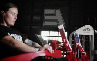 Person wearing earbuds sits on a red gym machine in a dim, industrial-style fitness space with a large A logo on the wall behind them.