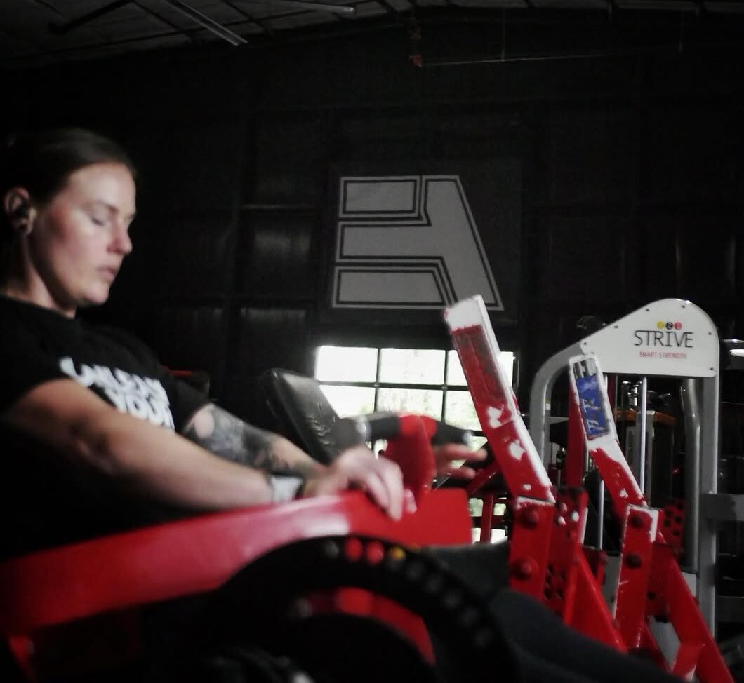 Person wearing earbuds sits on a red gym machine in a dim, industrial-style fitness space with a large A logo on the wall behind them.